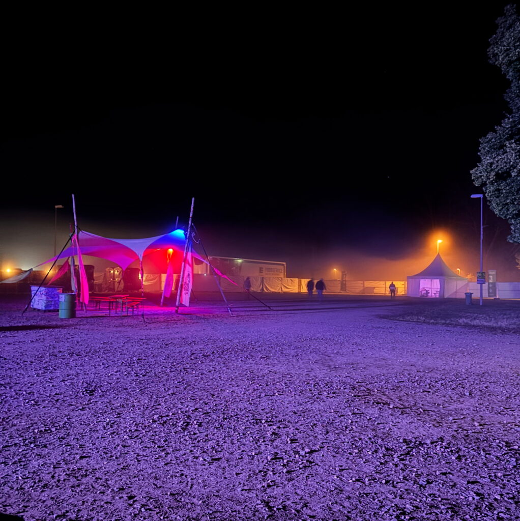 The Zappanale Festival ground on the closing night in 2025. There’s a tent with seating underneath, lit up in red and gold, with a black starlit sky above it. There’s mist in the air.